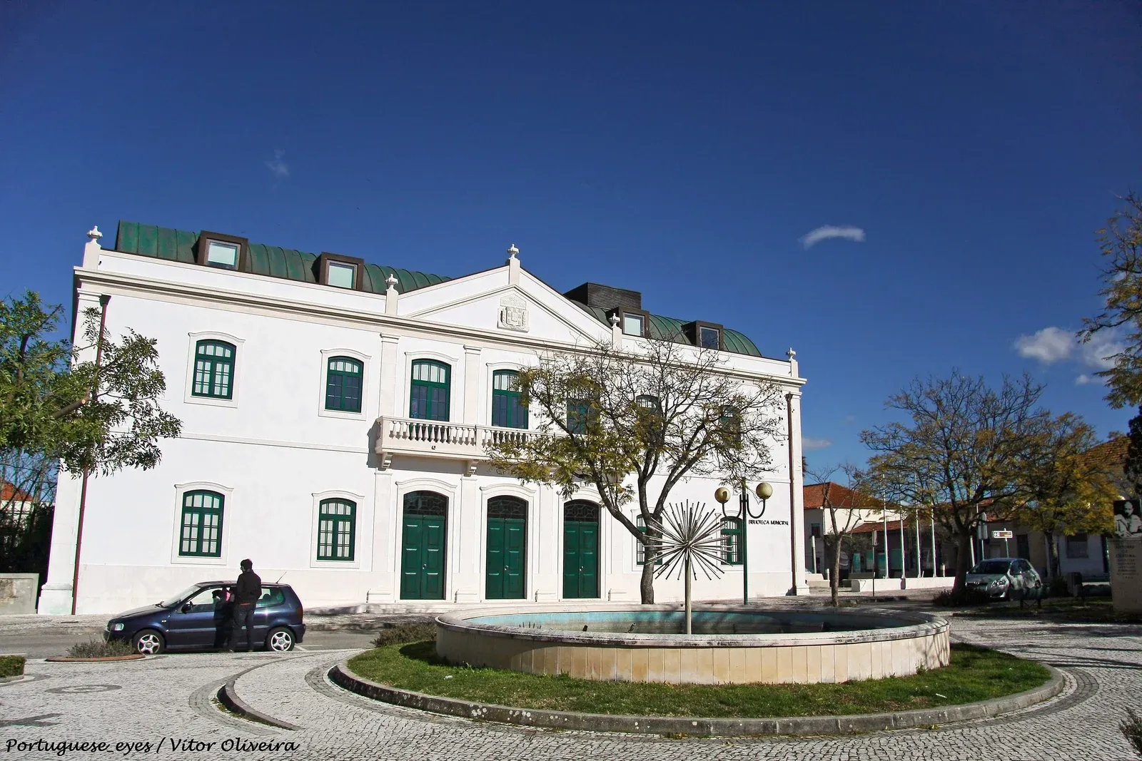 Biblioteca Municipal de Oliveira do Bairro, com fachada branca, janelas verdes e fonte na praça